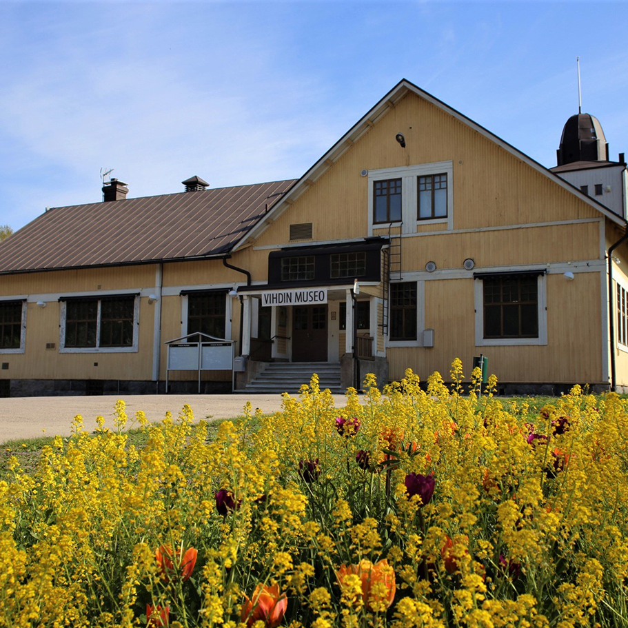 A flower garden in the foreground, a yellow Art Nouveau wooden building in the background.