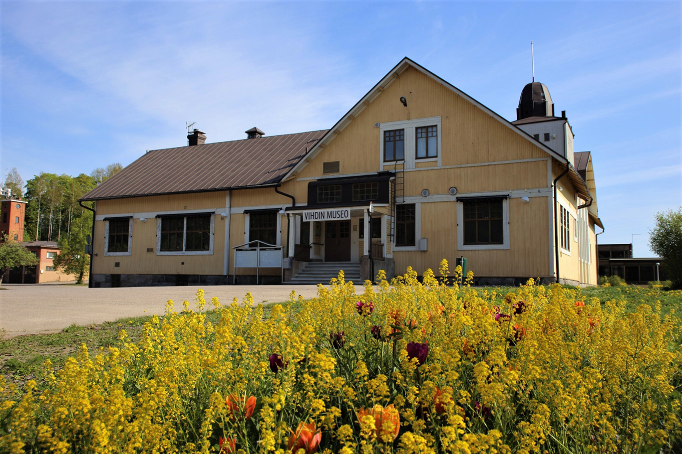 A flower garden in the foreground, a yellow Art Nouveau wooden building in the background.
