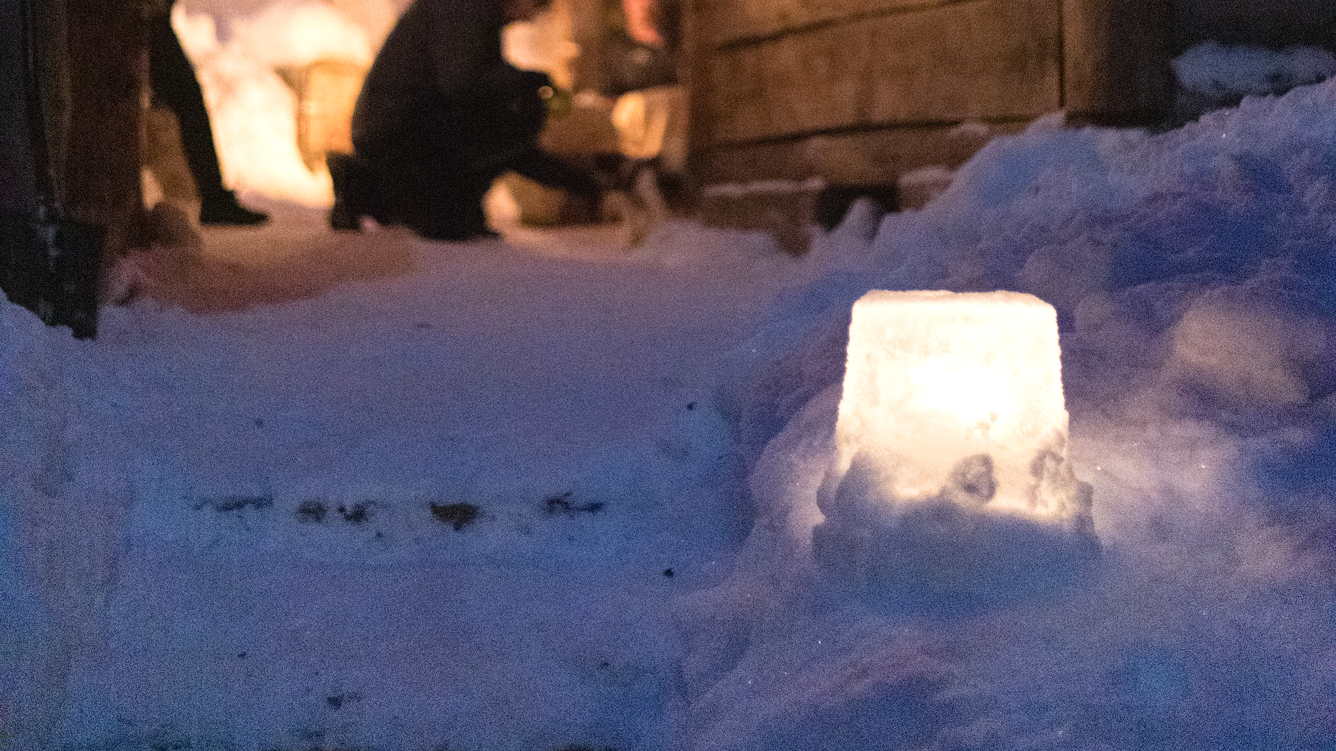 Lumilyhty, taka-alalla mökkirakennuksia ja henkilö sytyttämässä toista lumilyhtyä.